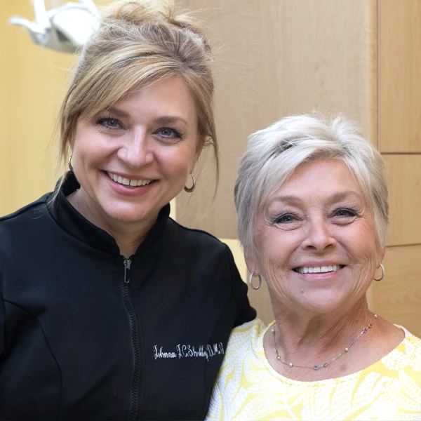 Johnna Shockley and her mother standing side by side in a clinical setting, facing the camera with shelving in the background.