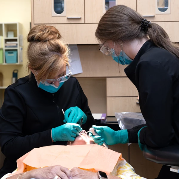 Two dental professionals wearing protective equipment performing an examination on a reclining patient.