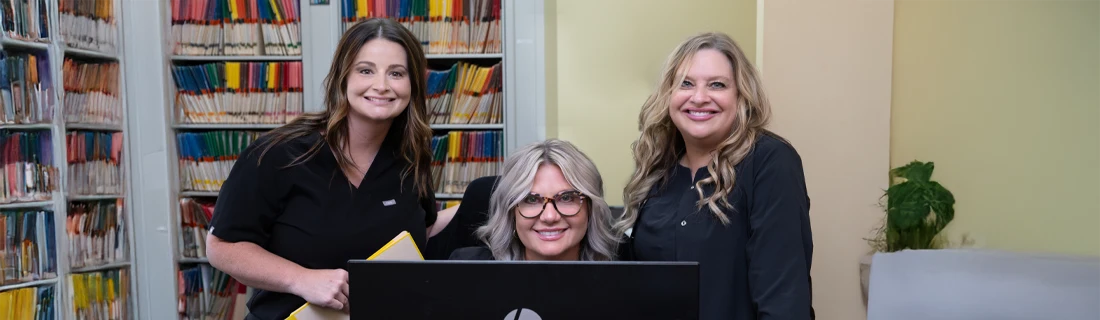 Three office staff members gathered around a computer workstation with file cabinets in the background.