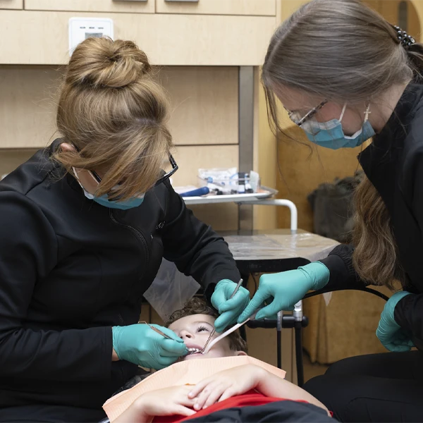 Two dental professionals wearing masks and gloves treating a child reclining in a dental chair