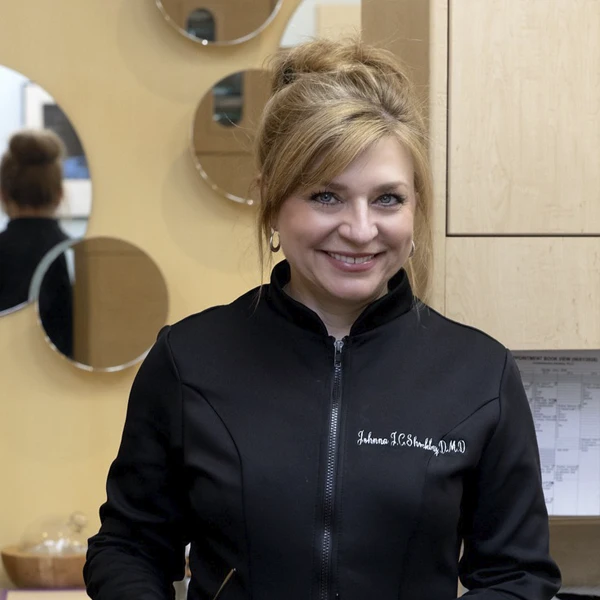 Portrait of a dental professional standing in a hallway wearing a black clinical jacket and glasses.